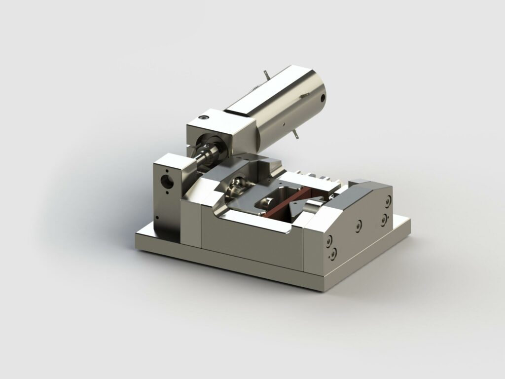 Auf einer flachen Basis sitzt ein metallisches Präzisionsinstrument mit verschiedenen mechanischen Komponenten. Das Gerät weist einen nach oben ragenden zylindrischen Teil und weitere mit Schrauben und Bolzen befestigte Teile auf, was auf eine spezielle Biege-Modul-Werkzeugmaschine oder ein Prüfgerät hindeutet.