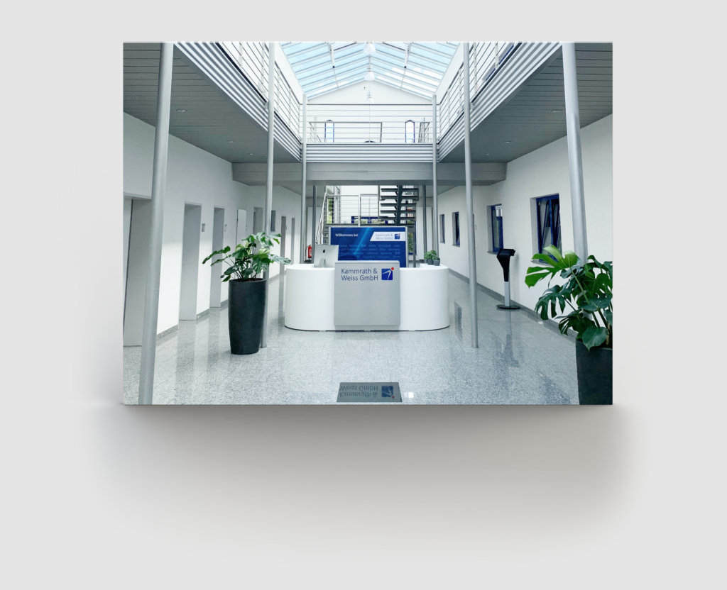 A clean, modern office lobby with a white reception desk at the center, flanked by two large potted plants. Sparse furniture lines the white walls. Overhead, natural light streams through large skylights. In the background, a staircase leads to an upper floor, inviting further contact and exploration.